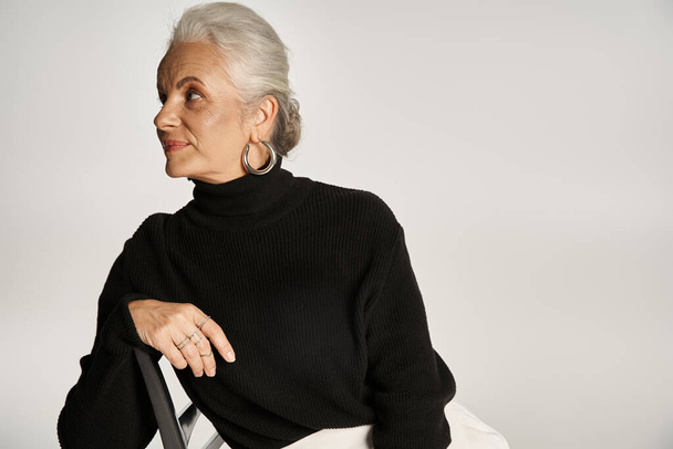 urocza kobieta w średnim wieku w eleganckim stroju casual i kolczyki obręczy siedzi na krześle na szarości - Zdjęcie, obraz