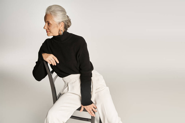 attraktive Frau mittleren Alters in schicker Freizeitkleidung und Reifrohrringen sitzt auf einem Stuhl auf grau - Foto, Bild