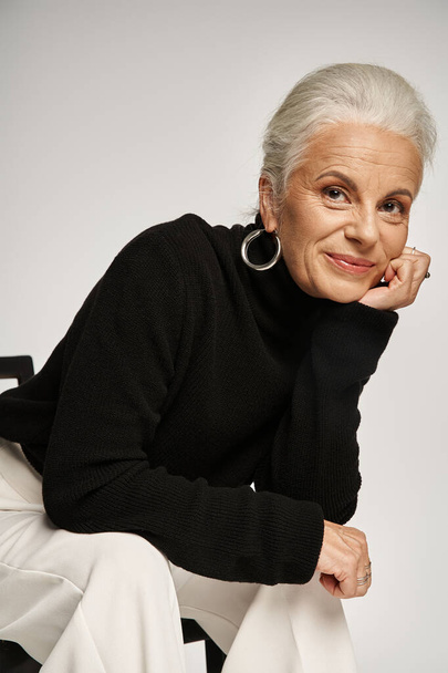 aantrekkelijke vrouw van middelbare leeftijd in slimme casual kleding en hoepel oorbellen zitten op stoel op grijs - Foto, afbeelding