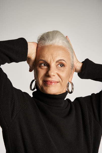 portret van gelukkige middelbare leeftijd zakenvrouw in elegante kledij en hoepel oorbellen op grijze achtergrond - Foto, afbeelding