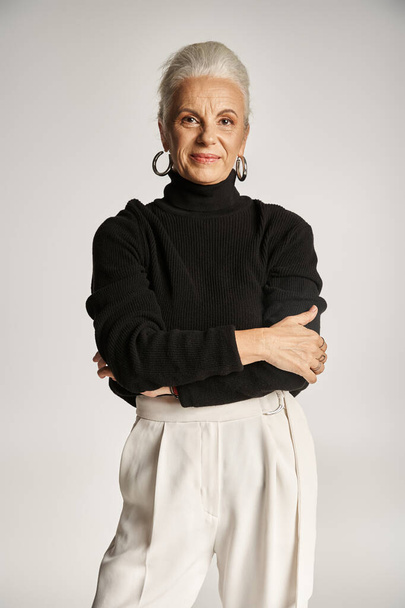 happy middle aged business woman in elegant attire and hoop earrings standing on grey backdrop - Photo, Image