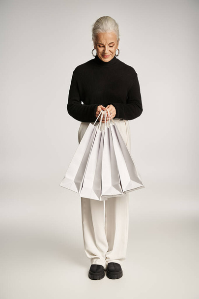 alegre mujer de mediana edad en traje elegante y pendientes de aro de pie con bolsas de compras en gris - Foto, Imagen