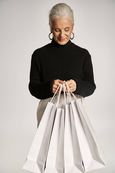 веселая женщина средних лет в элегантной одежде и серьги обруч стоя с пакетами для покупок на сером - Фото, изображение