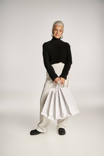 zadowolona kobieta w średnim wieku w eleganckim stroju i kolczykach obręczy stojących z torbami na zakupy na szaro - Zdjęcie, obraz