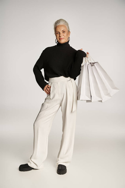 mujer de mediana edad satisfecha en elegante atuendo y pendientes de aro de pie con bolsas de compras en gris - Foto, imagen