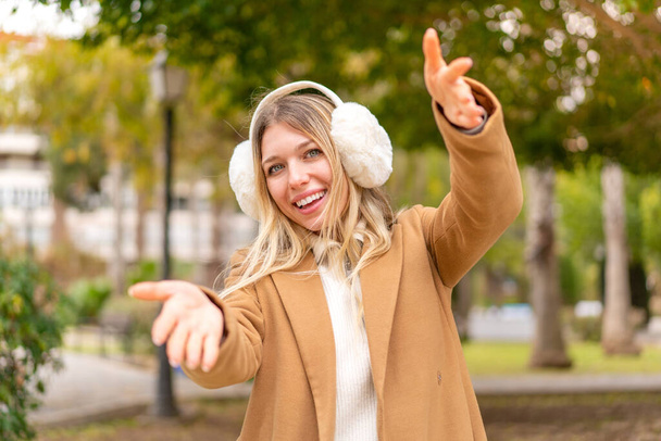 Молодая красивая блондинка в зимних муфтах на открытом воздухе представляет и приглашает прийти с рукой - Фото, изображение