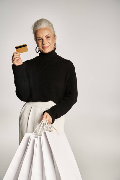 задоволена жінка середнього віку в елегантному вбранні тримає кредитну картку і сумки для покупок на сірому тлі - Фото, зображення