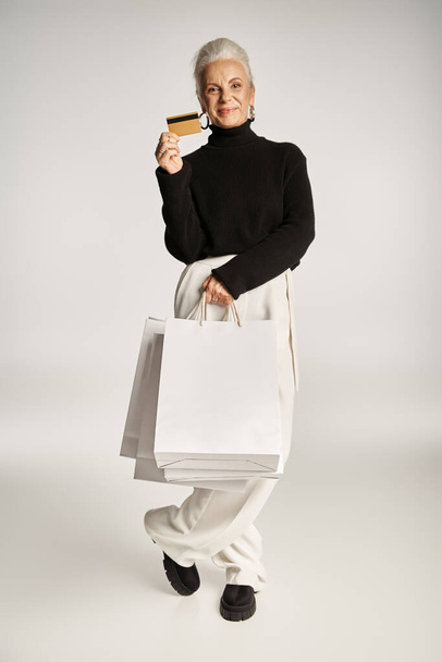 tevreden middelbare leeftijd vrouw in elegante kleding met creditcard en boodschappentassen op grijze achtergrond - Foto, afbeelding