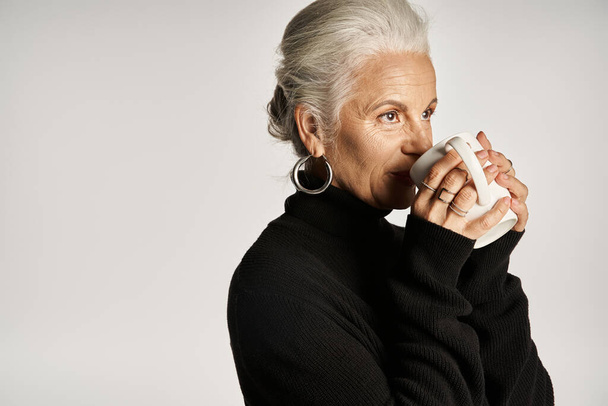 happy middle aged woman in black turtleneck sweater holding cup of coffee on grey backdrop - Photo, Image