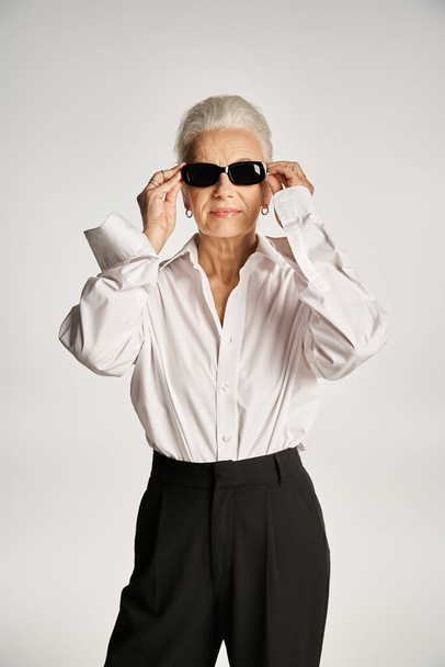 élégante femme d'âge moyen avec des cheveux gris portant des lunettes de soleil et debout en tenue élégante sur gris - Photo, image