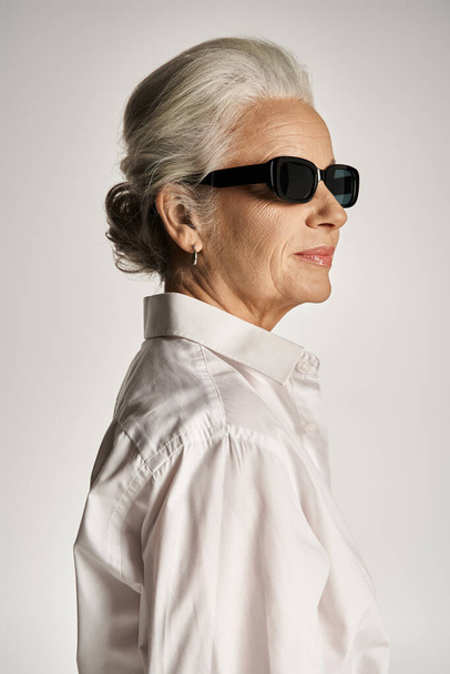 portrait de femme élégante d'âge moyen en chemise blanche et lunettes de soleil posant sur fond gris - Photo, image