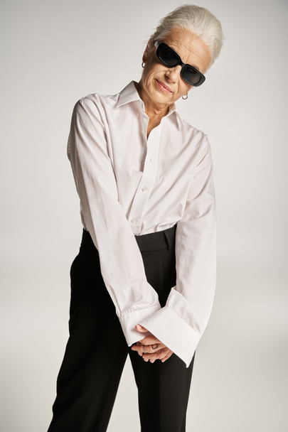 alla moda donna di mezza età in camicia bianca, pantaloni neri e occhiali da sole in posa su sfondo grigio - Foto, immagini