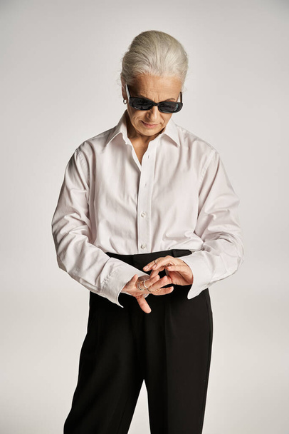 élégante femme d'âge moyen en chemise blanche, pantalon noir et lunettes de soleil regardant des anneaux sur les doigts - Photo, image