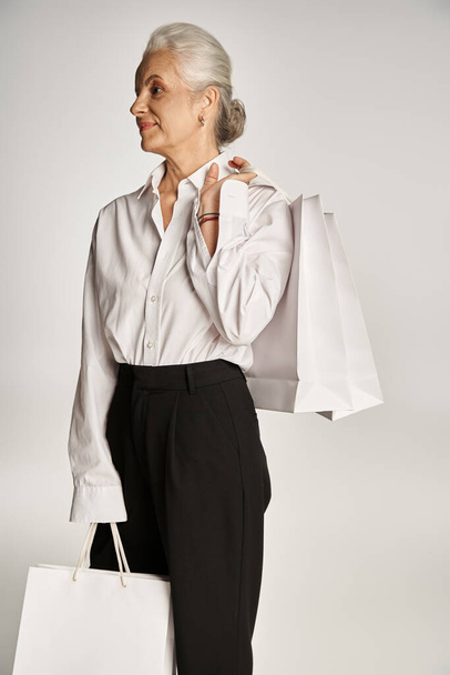 stylowa kobieta w średnim wieku w eleganckiej białej koszuli i czarne spodnie stojące z torbami na zakupy na szary - Zdjęcie, obraz
