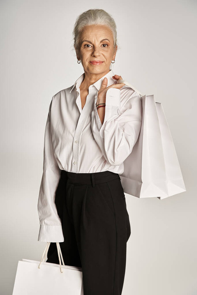 élégante femme d'âge moyen en chemise blanche chic et pantalon noir debout avec des sacs à provisions sur gris - Photo, image