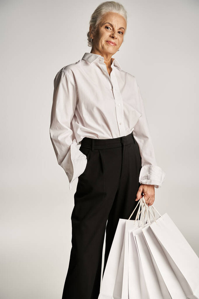 mujer de mediana edad en camisa blanca y pantalones de pie con la mano en el bolsillo y la celebración de bolsas de compras - Foto, Imagen