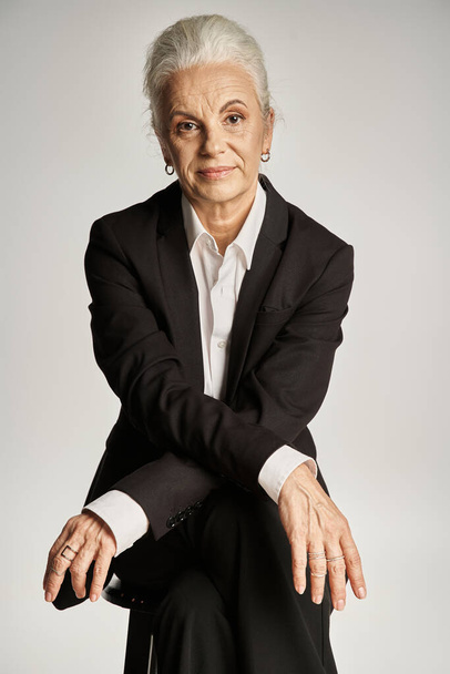 mooie en grijze harige vrouw van middelbare leeftijd in elegante kleding rusten op grijze achtergrond - Foto, afbeelding
