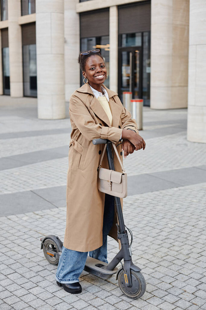 Retrato de comprimento total vertical de jovem mulher negra montando scooter elétrico na cidade e sorrindo alegremente para a câmera enquanto vestindo casaco longo - Foto, Imagem