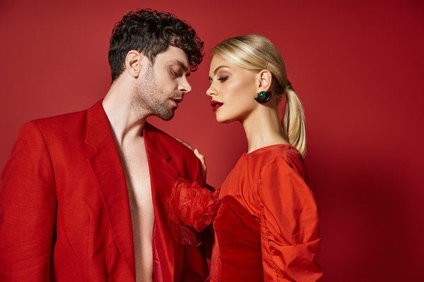 fashionable couple in vibrant attire standing together on red background, man and woman in red - Photo, Image