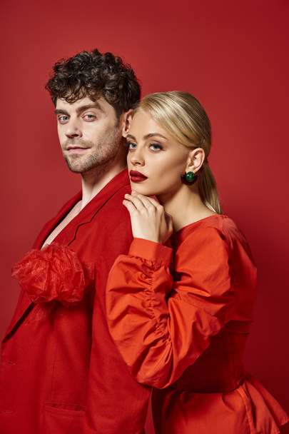 blonde woman with red lips leaning on shoulder of man in vibrant attire of red background, fashion - Photo, Image