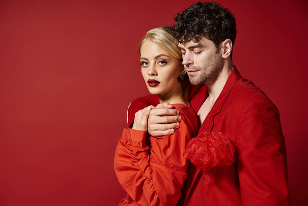 schöner Mann umarmt blonde Frau in eleganter Kleidung auf rotem Hintergrund, Mode - Foto, Bild