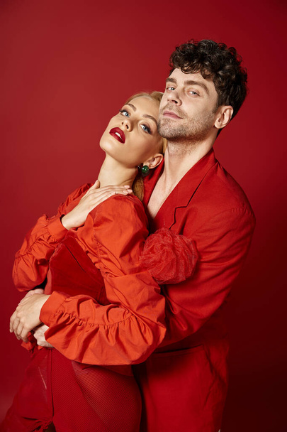 handsome man embracing beautiful woman in elegant attire on red background, stylish couple - Фото, зображення