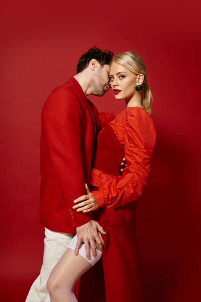 schöner Mann berührt Bein der verführerischen blonden Frau in sexy Strümpfen auf rotem Hintergrund, Mode - Foto, Bild