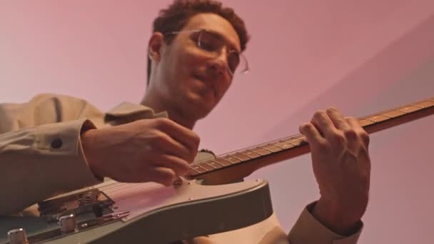 Disparo de bajo ángulo de músico masculino tocando la guitarra y sonriendo en el estudio de casa con luz de neón - Metraje, vídeo