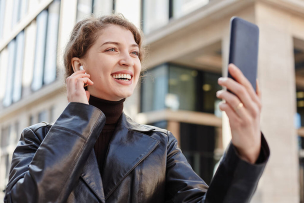 Retrato de una joven sonriente llamando por video chat en la ciudad y ajustando auriculares inalámbricos - Foto, Imagen
