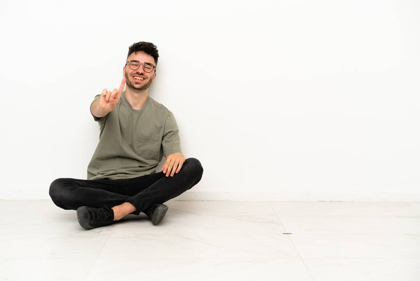 Jovem caucasiano sentado no chão isolado no fundo branco mostrando e levantando um dedo - Foto, Imagem