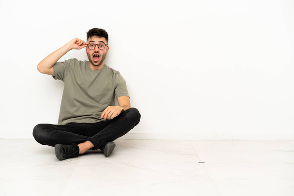Joven hombre caucásico sentado en el suelo aislado sobre fondo blanco con gafas y sorprendido - Foto, Imagen