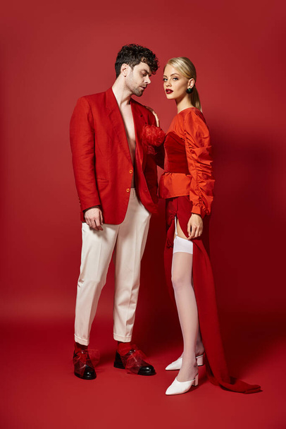 pohledný muž v červené značkové oblečení pózování s blondýnou modelu na pulzujícím pozadí, módní pár - Fotografie, Obrázek