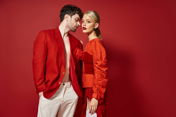 hombre guapo en traje de diseñador rojo posando con modelo rubio sobre un fondo vibrante, pareja de moda - Foto, Imagen