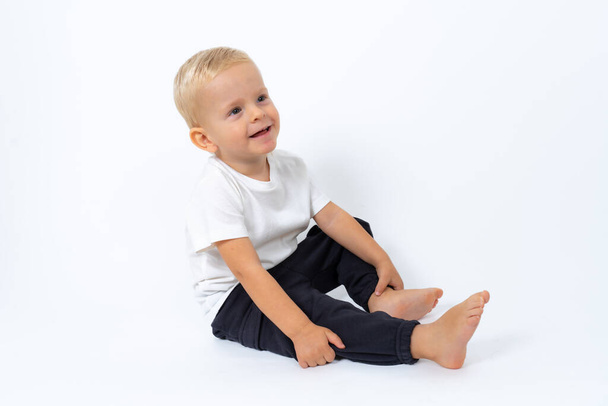blondyn chłopiec w białym t-shirt i czarne spodnie siedzi na podłodze i pozowanie na białym tle studio - Zdjęcie, obraz