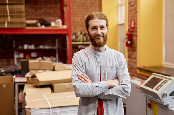 Porträt eines lächelnden jungen Mannes, der in einer industriellen Druckerei arbeitet und mit verschränkten Armen steht, Kopierraum - Foto, Bild