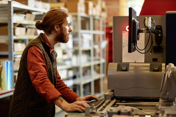 Πλευρική άποψη πορτρέτο του αρσενικού εργαζομένου που χειρίζεται εκτυπωτική μηχανή στο εργοστάσιο εκτύπωσης - Φωτογραφία, εικόνα