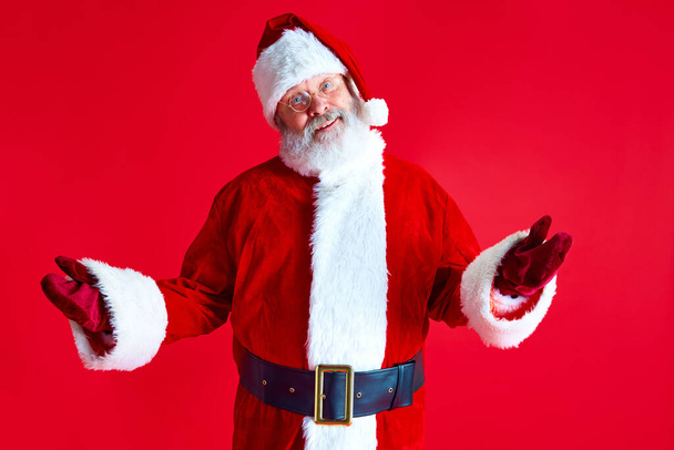 Mature man with long beard, kind eyes dressed as Santa with open arms accepts Happy New Year wishes against red studio background. Concept of winter holidays, Merry Christmas, joy and fun. - Photo, Image