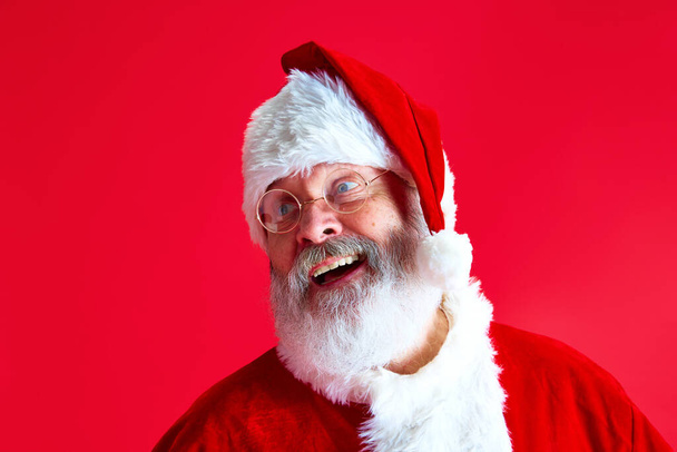 Close-up portret van volwassen man met lange grijze baard in glazen gekleed in Santa Claus feestelijke kostuum vrolijk glimlachen tegen rode achtergrond. Concept van de wintervakantie, Vrolijk kerstfeest, vreugde en plezier - Foto, afbeelding