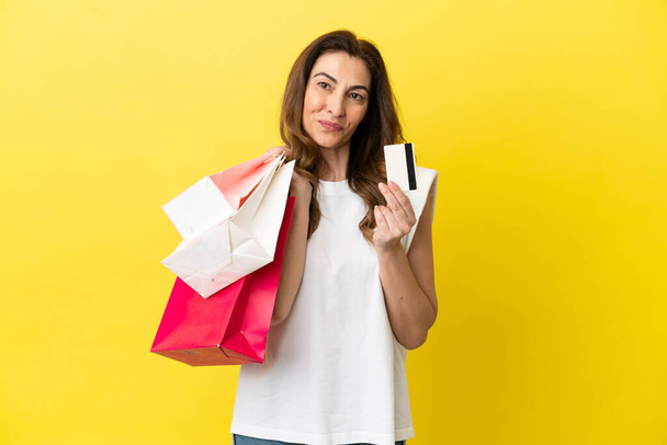 Mujer caucásica de mediana edad aislada sobre fondo amarillo sosteniendo bolsas de compras y una tarjeta de crédito - Foto, Imagen