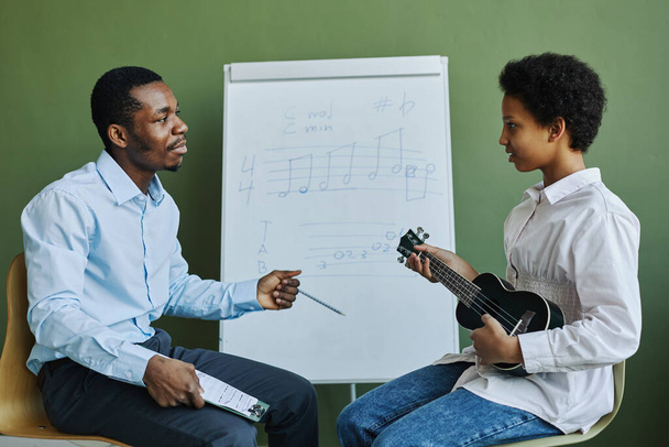 młody pewny siebie afro-amerykański nauczyciel odpowiadając na pytania uczennicy na lekcji z ukulele podczas obu siedzi przez tablicę - Zdjęcie, obraz