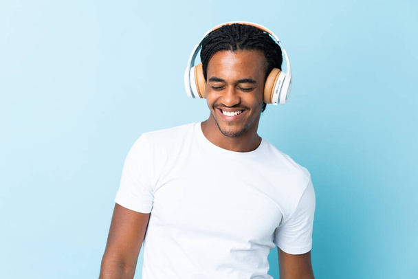 Jonge Afro-Amerikaanse man met vlechten geïsoleerd op blauwe achtergrond luisteren muziek - Foto, afbeelding