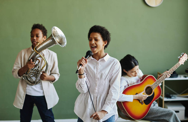 Щаслива молода школярка з мікрофоном виконує пісню з двома однокласниками, які грають на трубі та акустичній гітарі на уроці - Фото, зображення
