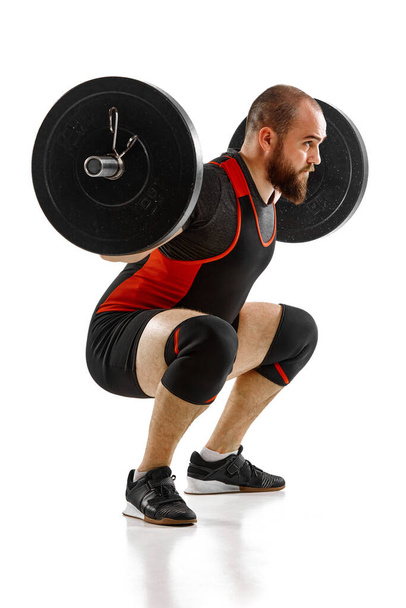Strong, muscular, bearded man, bodybuilder doing squats and lifting barbell, heavy weights against white background. Side view. Concept of sport, strength, gym, healthy lifestyle, power, weightlifting - Fotografie, Obrázek