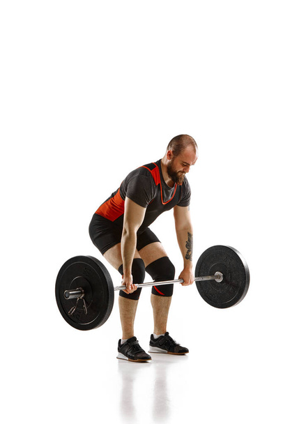 Bearded, muscular, young man, weightlifter training, doing exercise of deadlift with barbell over white background. Concept of sport, strength, gym, healthy lifestyle, power, endurance, weightlifting - 写真・画像