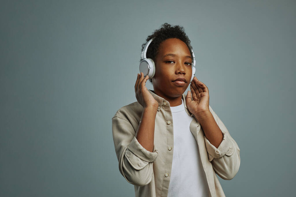 Νεανικό σχολιαρόπαιδο που αγγίζει λευκά ακουστικά ακούγοντας την αγαπημένη του μουσική και κοιτάζοντας την κάμερα πάνω από γκρι φόντο - Φωτογραφία, εικόνα