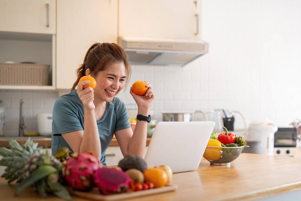 Šťastná žena asijské ve stylové teplákové soupravě čerstvé smoothie natáčení videa s moderním notebookem v kuchyni. - Fotografie, Obrázek
