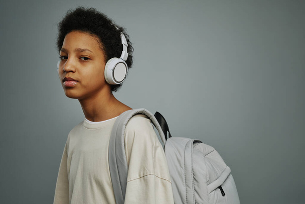 Colegiala bastante joven con mochila de pie delante de la cámara y escuchar música en los auriculares mientras va a la escuela - Foto, Imagen