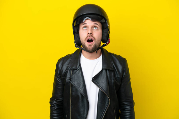 Mladý běloch s motocyklovou helmou izolované na žlutém pozadí vzhlíží a s překvapeným výrazem - Fotografie, Obrázek