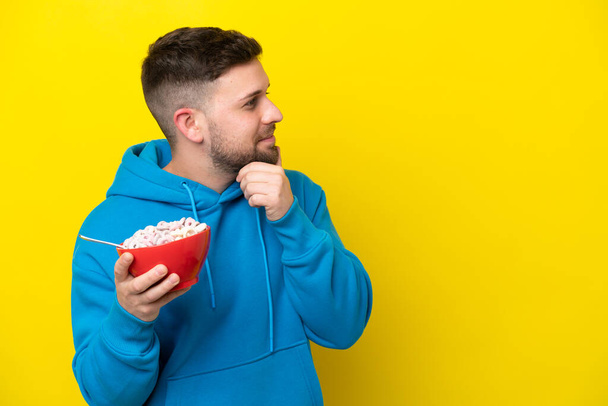 Joven hombre caucásico sosteniendo un tazón de cereales aislado sobre fondo amarillo pensando en una idea y mirando al lado - Foto, Imagen