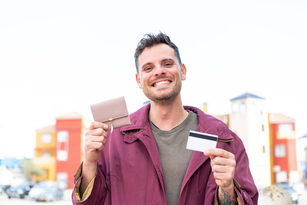 Молодий білий чоловік на відкритому повітрі тримає гаманець і кредитну картку - Фото, зображення